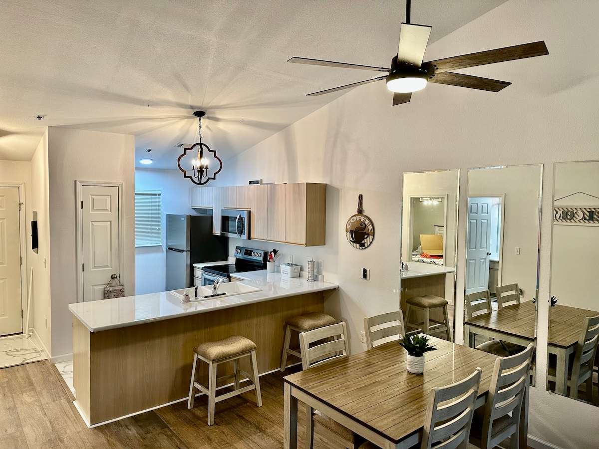An open kitchen area is featured, showcasing modern appliances and a spacious island with seating for three. A wooden dining table accommodates four, with decorative mirrors reflecting the light from a ceiling fan and stylish light fixtures.
