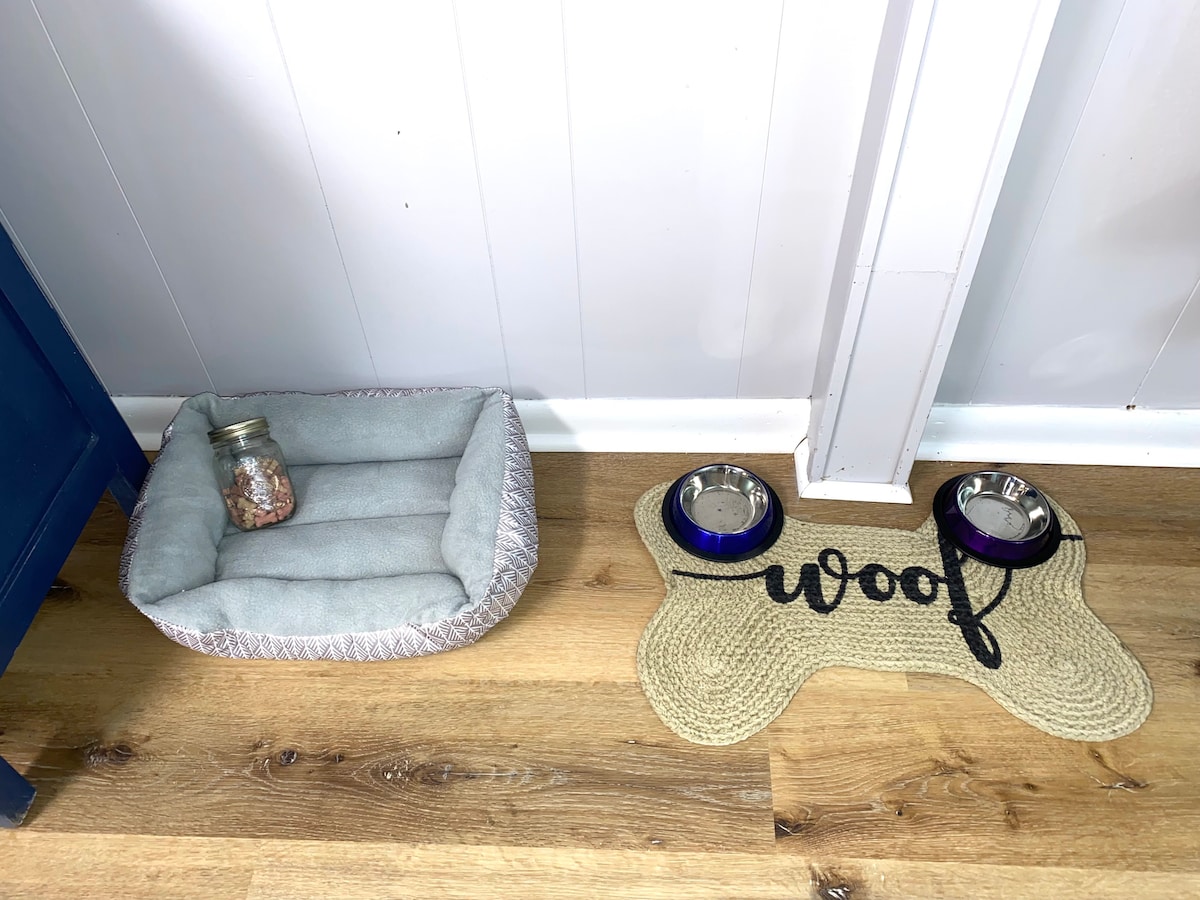 A designated pet area is displayed, featuring a soft gray pet bed alongside a woven mat with 'woof' text. Two metal bowls are positioned on the mat, providing a comfortable space for pets.