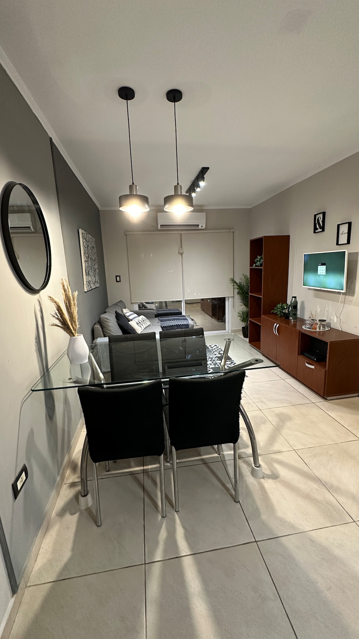 A modern living space features a glass dining table paired with black chairs, illuminated by pendant lights overhead. A comfortable sofa is positioned nearby, with decorative accents adding character. A television is mounted on the wall opposite a contemporary cabinet, creating a functional and inviting area.
