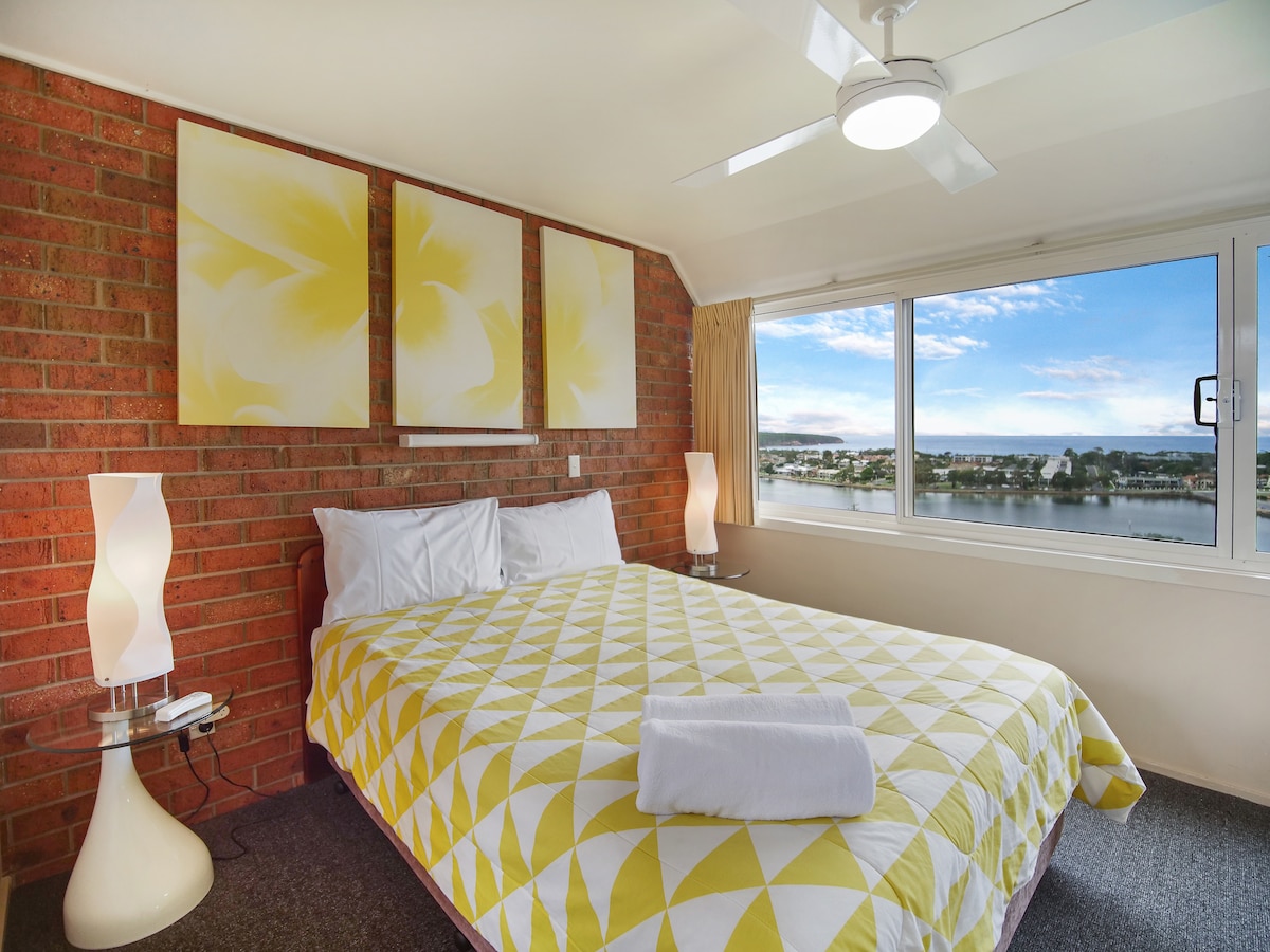 A bright bedroom features a yellow and white patterned quilt atop a spacious bed. Two bedside lamps illuminate the room, and large windows provide a view of the waterfront. Three abstract art pieces in yellow hang above the bed, complementing the brick accent wall.