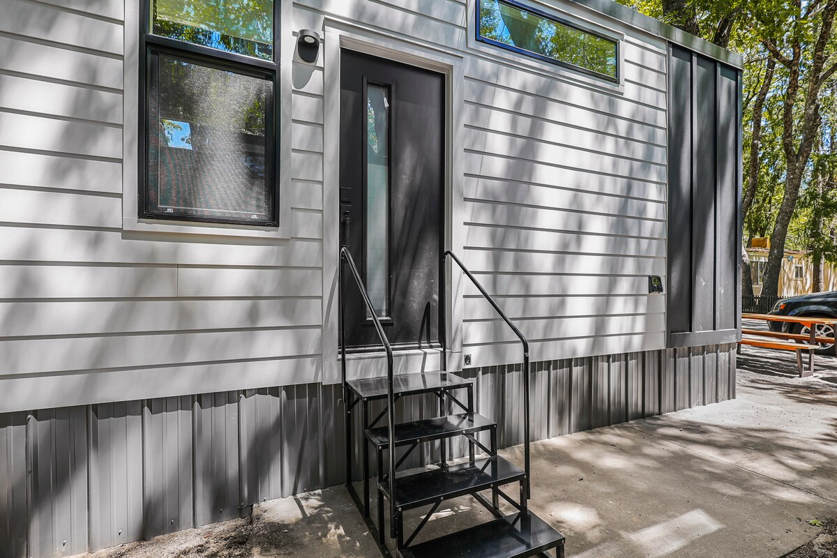 The entrance is framed by a modern exterior featuring white paneling and large windows that allow natural light to filter in. A set of black metal stairs leads to the door, enhancing accessibility. Nearby, a picnic area with a long wooden table can be seen.
