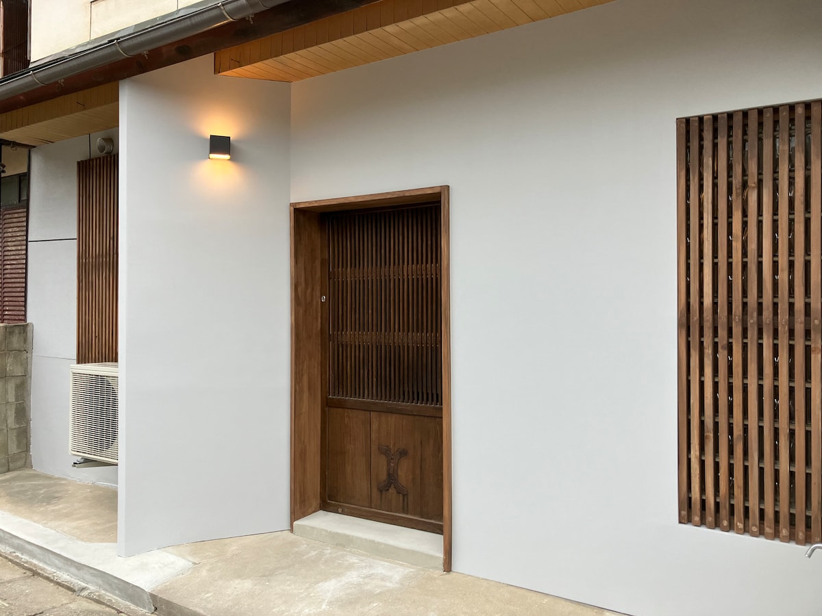 The entrance to the traditional guesthouse features a wooden door with horizontal slats, providing glimpses of the interior. A modern light fixture above the door complements the clean, white walls. The exterior also includes wooden lattice windows on either side.