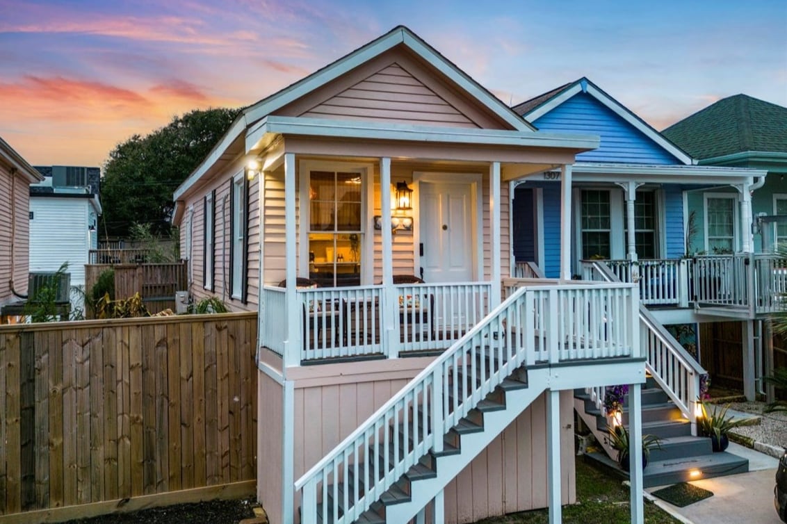 A charming coastal cottage exterior is presented, featuring pastel pink siding and a welcoming front porch. Stairs lead to the entryway, which is framed by light fixtures. Adjacent homes in complementary colors create a cohesive neighborhood aesthetic, set against a colorful sunset sky.