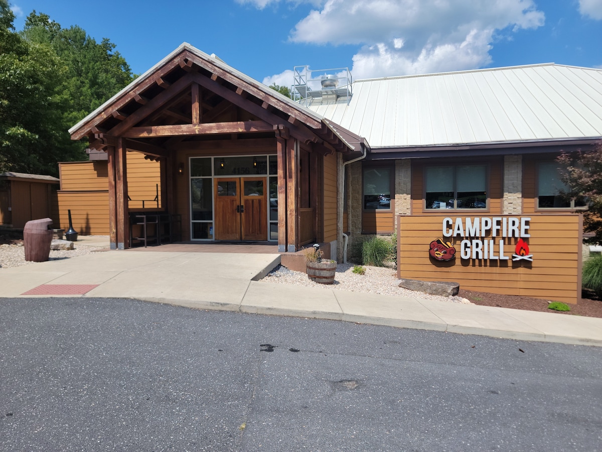A wooden entrance leads to the Campfire Grill, featuring a rustic design with a sloped roof and large windows. The walkway is lined with decorative stones and greenery, while a sign displaying the restaurant's name is prominently mounted on the exterior.