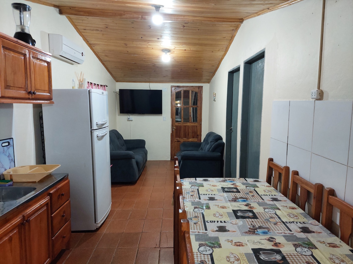 A well-equipped kitchen area features wood cabinetry and a refrigerator. A dining table with patterned tablecloth sits nearby. A comfortable living space is provided with two upholstered sofas facing a wall-mounted television. The room is illuminated by overhead lighting with wooden ceiling accents.
