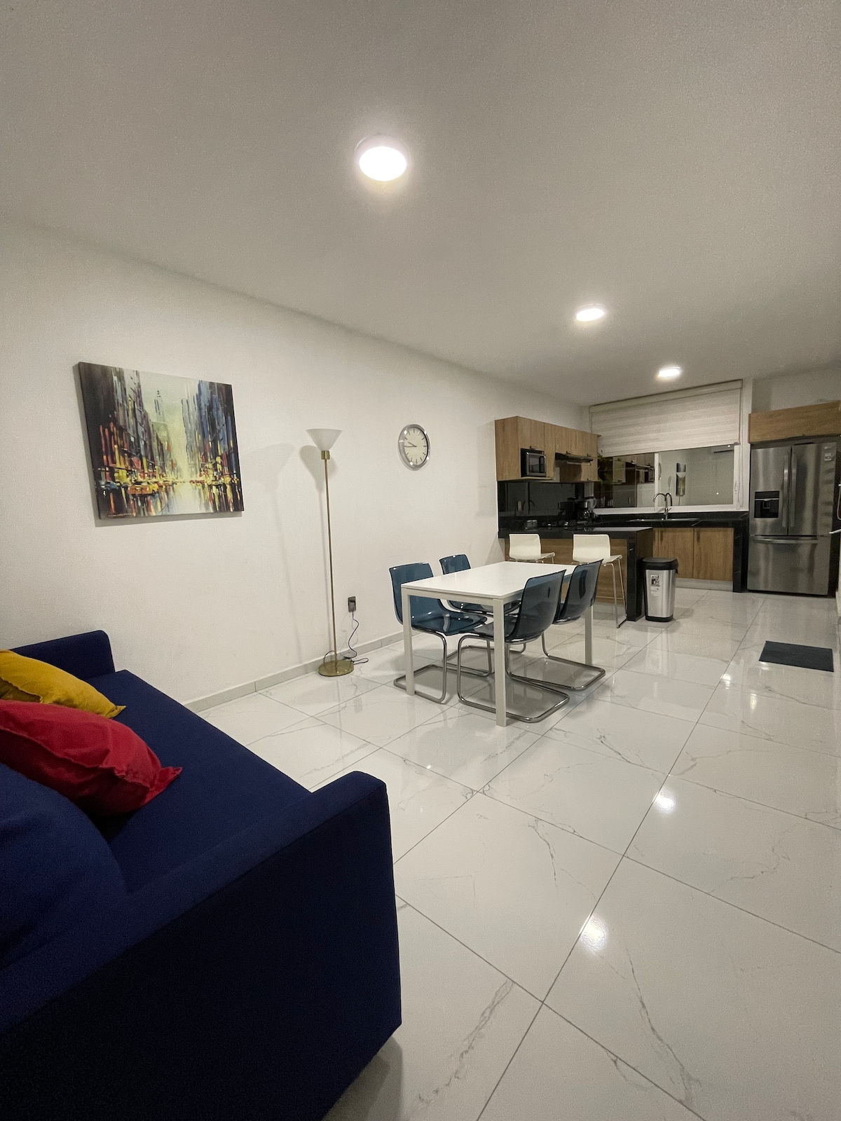 A modern living area is visible, featuring a blue sofa adorned with colorful cushions. A dining table with blue chairs is positioned in front of a well-equipped kitchen, which includes light wood cabinetry and stainless steel appliances. Bright overhead lights illuminate the space.