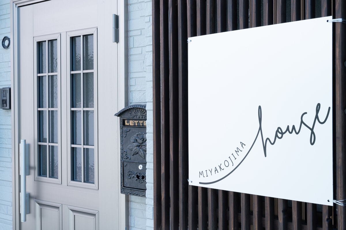 The entrance features a double door with multiple small window panes, offering a glimpse into the interior. Beside the door, a vintage-style mailbox is mounted on the wall. A white sign with elegant lettering indicates the name of the house.