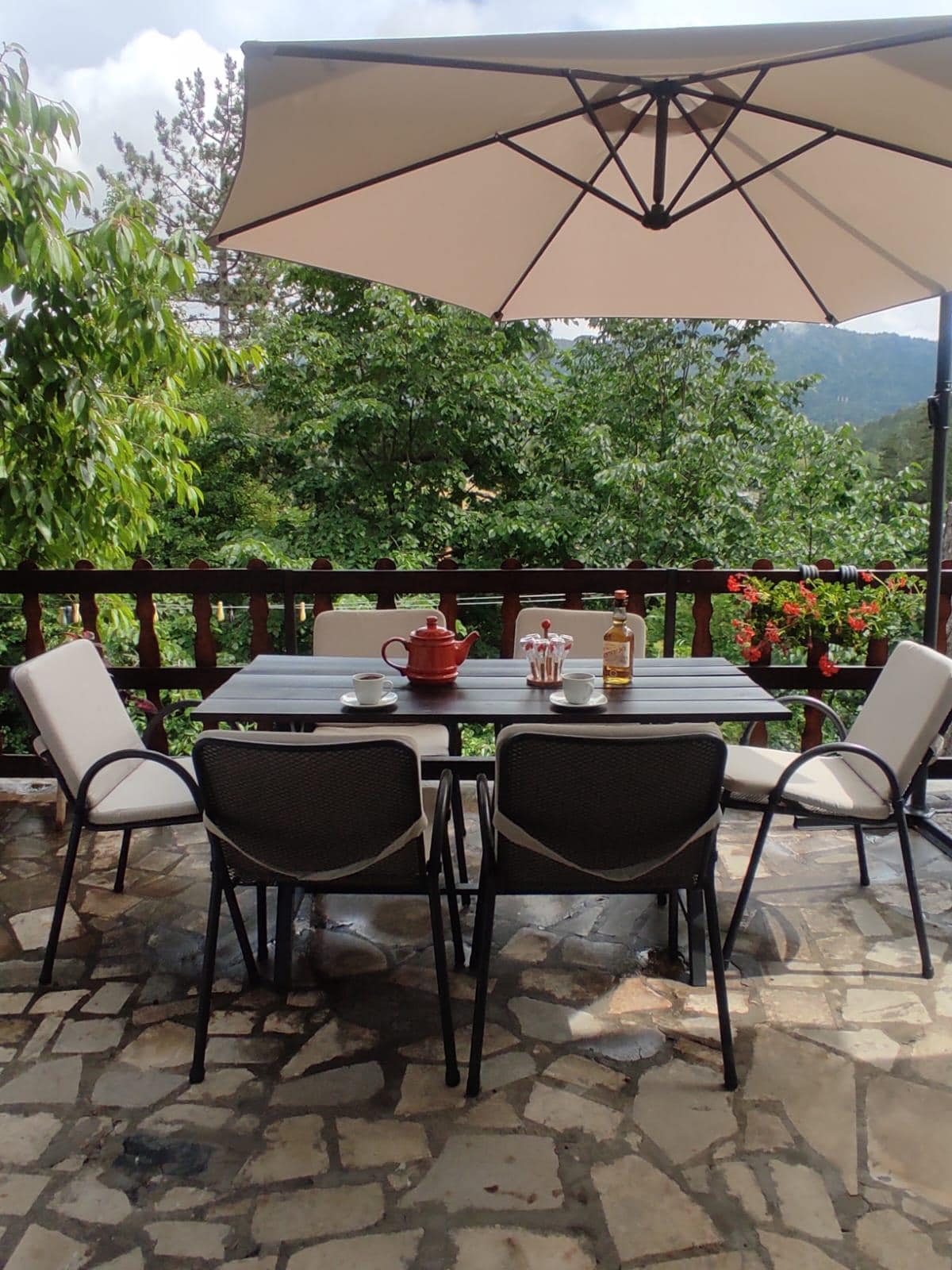 A covered terrace features a five-piece dining set with four chairs and a central table, surrounded by greenery. A large umbrella provides shade, while potted flowers add color. The flooring is composed of natural stone tiles, enhancing the outdoor dining experience.