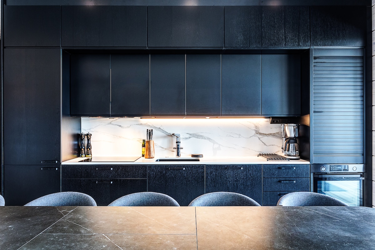 A contemporary kitchen features sleek black cabinetry and a stylish white marble backsplash. A fully equipped setup includes a coffee maker and stovetop, with ample countertop space for meal preparation. The dining table, positioned in front, is made of dark stone with rounded chairs.