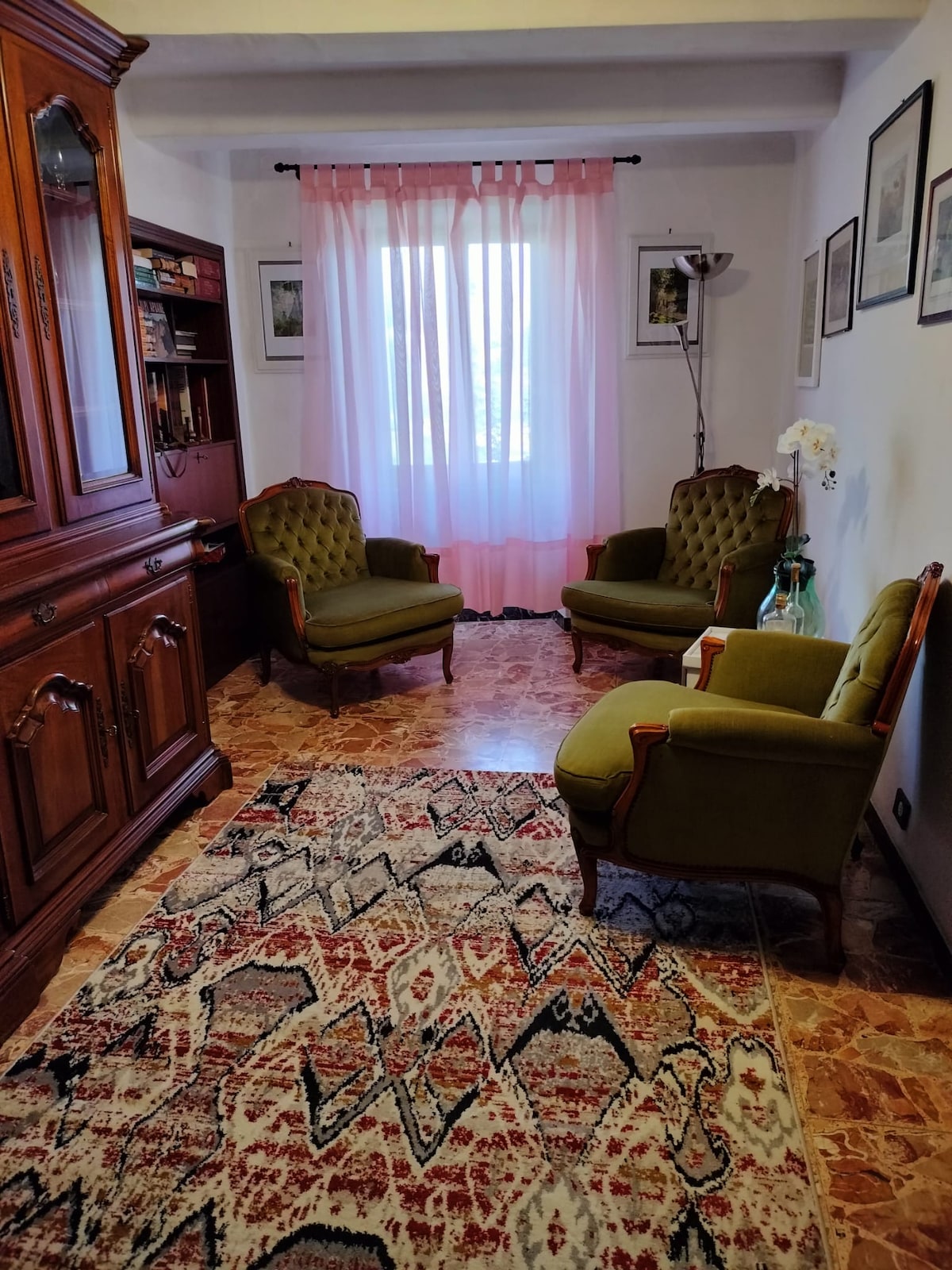 A welcoming sitting area features two upholstered green armchairs, positioned on a patterned rug. The space is brightened by a large window adorned with sheer pink curtains, complemented by framed artwork on the walls and a floor lamp to enhance the overall decor.