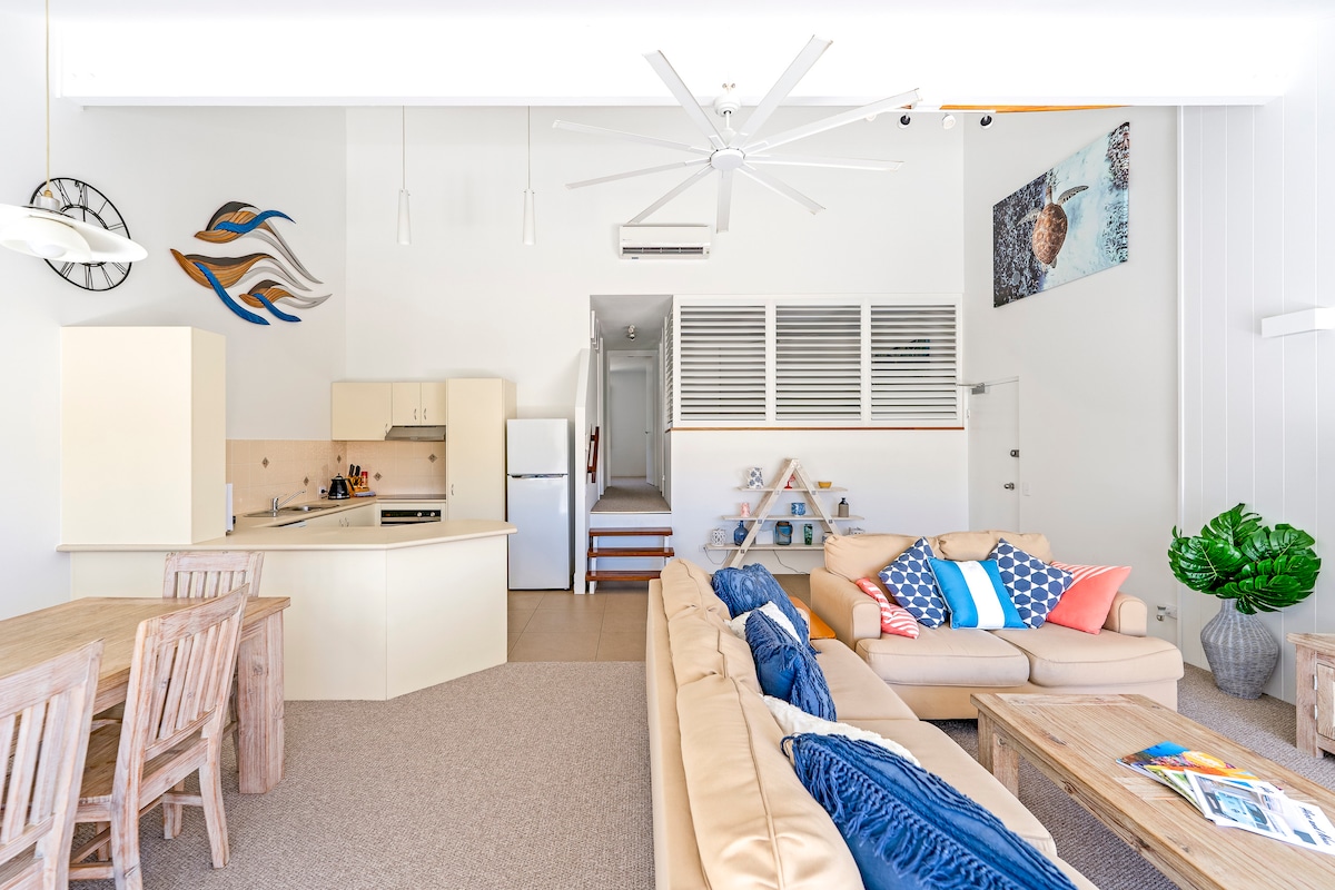 An open-plan living area showcases a light-coloured sofa adorned with cushions in various shades. A dining table crafted from wood is positioned nearby. A fully equipped kitchen features modern appliances, while decorative wall art enhances the ambience. Natural light floods the space through large windows.