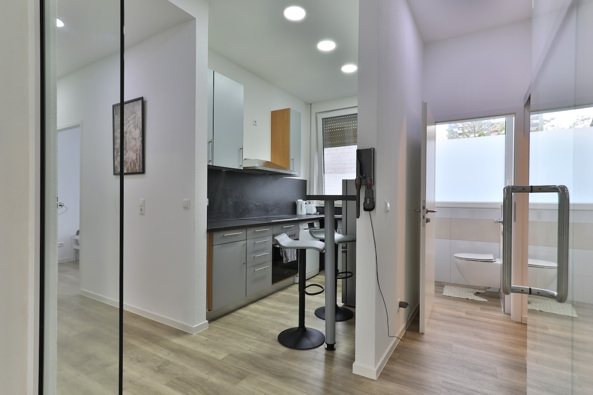 A modern kitchen is showcased, featuring sleek cabinetry, a dark countertop, and a set of two barstools. The open layout extends towards a well-lit space with large windows, providing a view into a bathroom area that includes a toilet and sink.