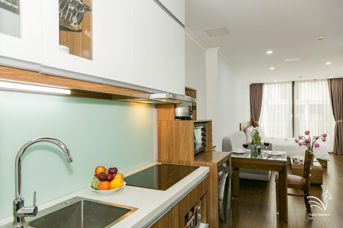 A well-equipped kitchen area showcases modern appliances, including a stove and oven. A decorative bowl of fresh fruit is positioned on the counter next to a dining table with chairs. Natural light floods the inviting living space through large windows adorned with light curtains.