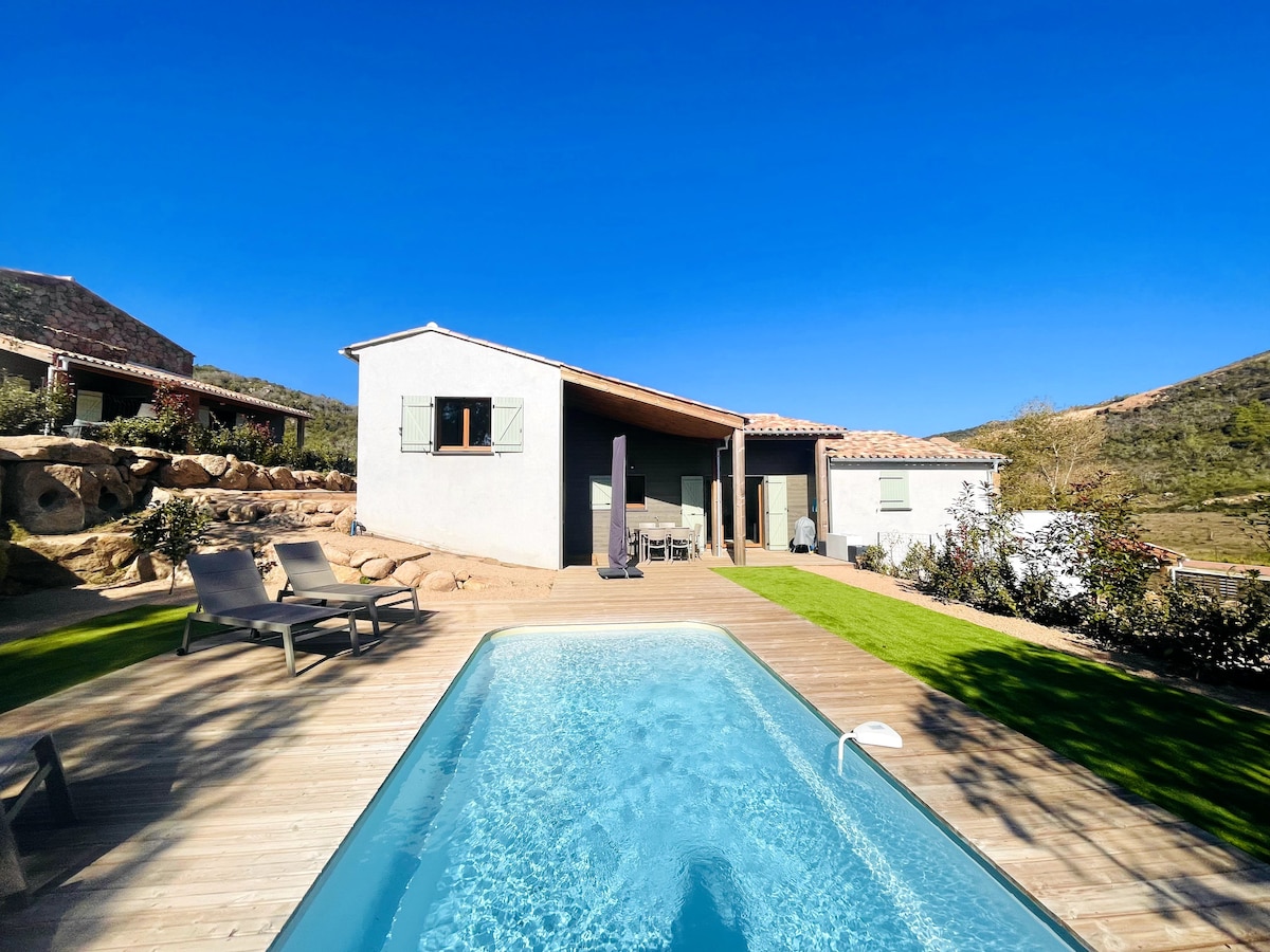 A private pool is framed by a wooden deck, showcasing lounging chairs arranged neatly beside it. The single-story house features multiple windows, a covered porch, and a well-maintained lawn, offering a calm environment with a clear blue sky above.