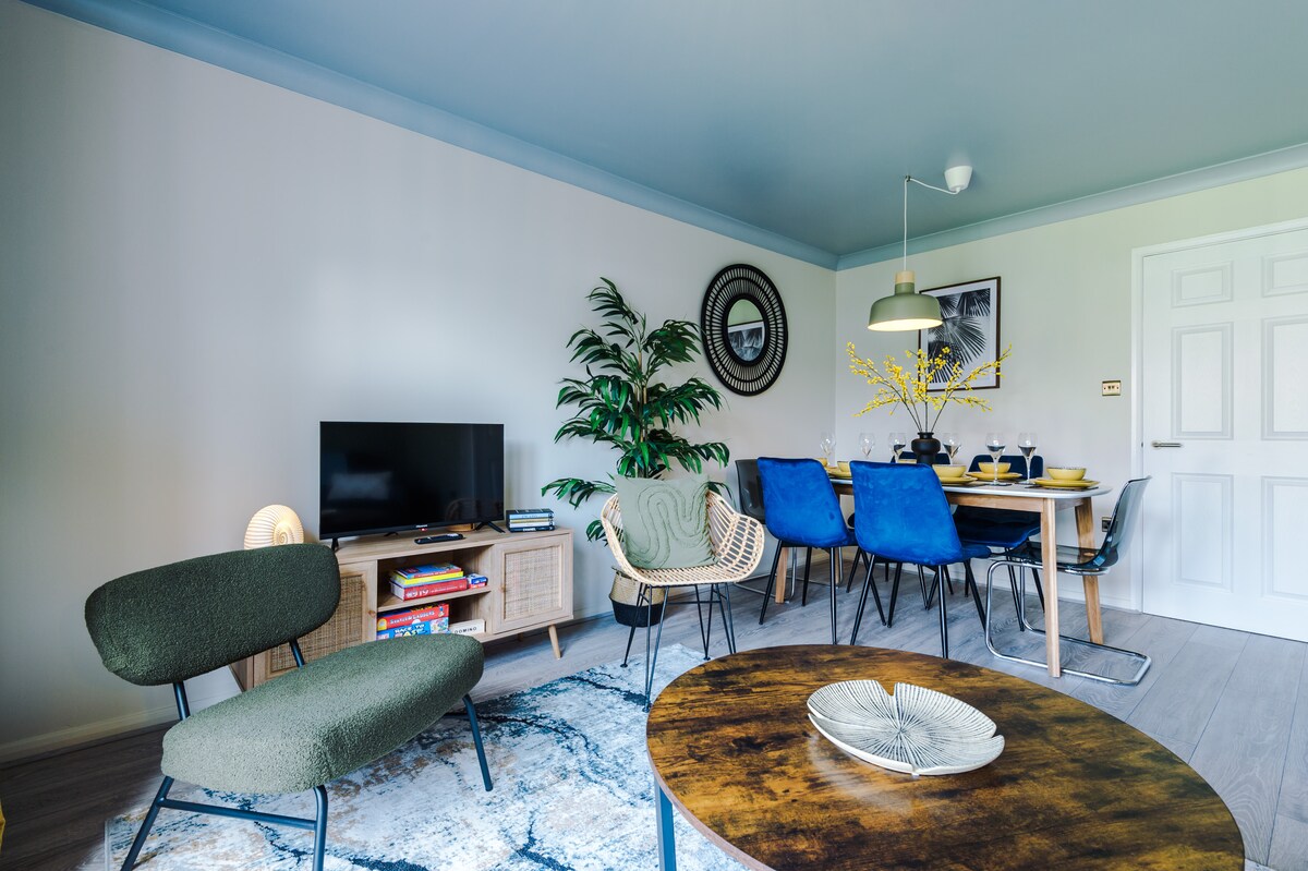 A comfortable living and dining area is presented, featuring a blend of modern furnishings. A green accent chair and a round wooden coffee table are included, alongside a television on a stylish console. The dining table is set with dishes, complemented by vibrant blue upholstered chairs.