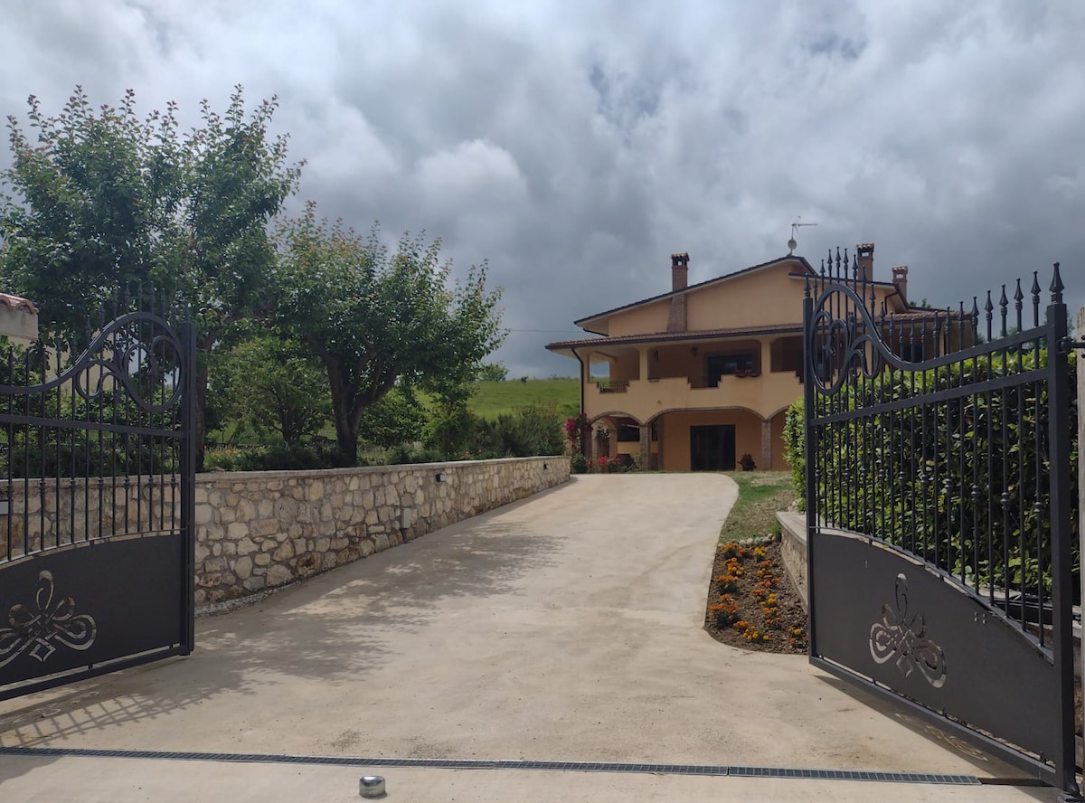 The entrance gate opens to a spacious driveway leading to a charming villa. Lush greenery flanks the sides, and the property features a two-story structure with a welcoming façade. Cloudy skies provide a dramatic backdrop to the inviting atmosphere.