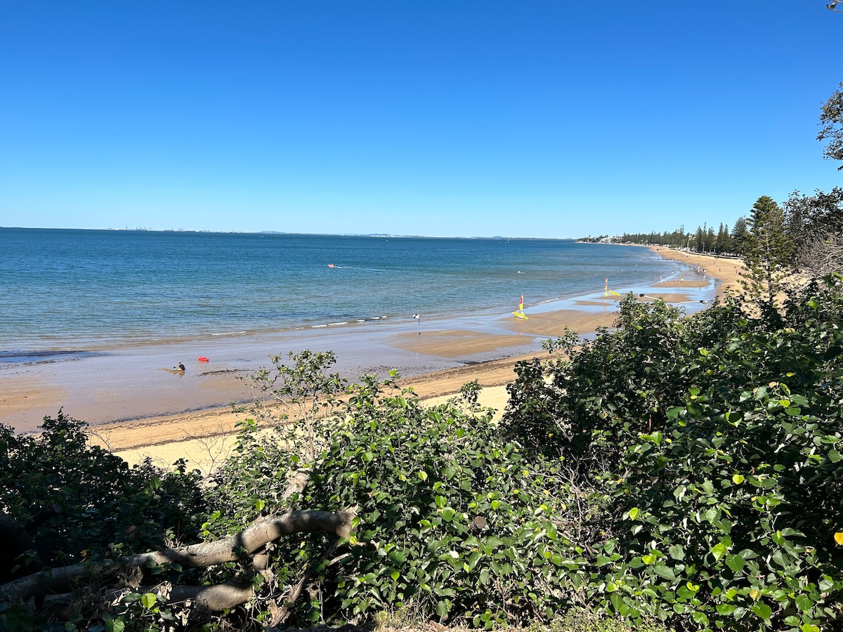 A serene beach scene is captured, featuring fine golden sand meeting calm blue waters under a clear blue sky. Sparse greenery frames the foreground, while a wide expanse of the shoreline stretches into the distance, where people can be seen enjoying the beach.