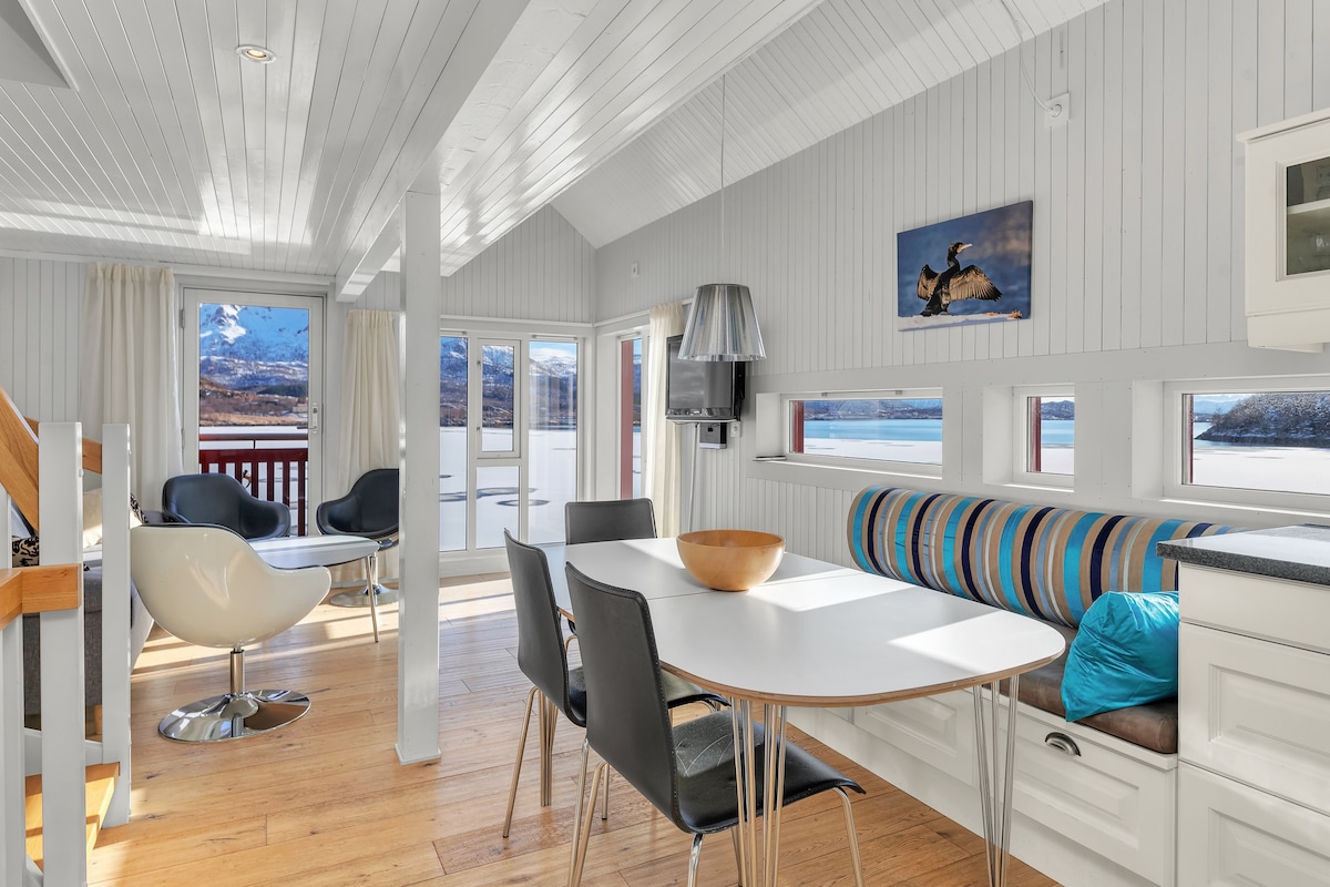 An airy dining area is featured with a white table surrounded by black chairs. A built-in seating area lines the wall, enhanced by colorful cushions. Large windows allow natural light to fill the space, showcasing views of the surrounding landscape.