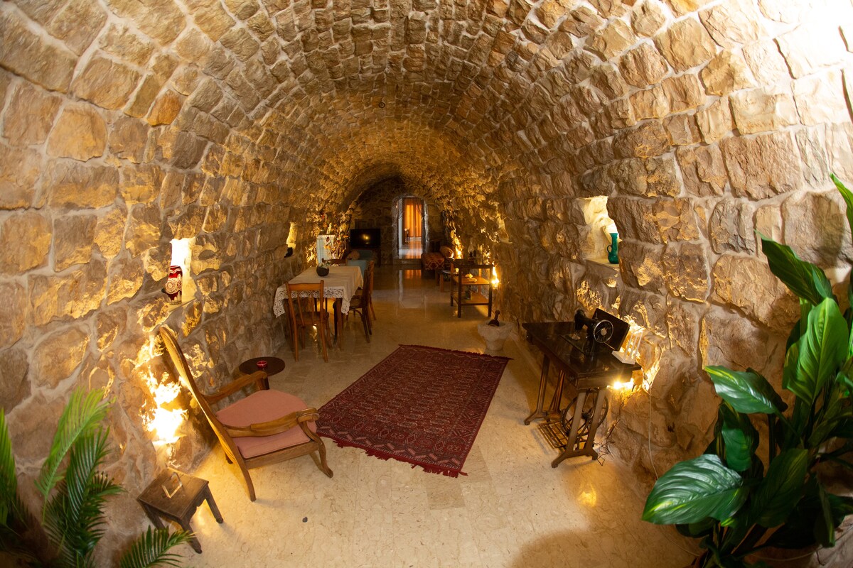 An arched stone hallway is illuminated with warm lighting, showcasing natural stone walls and a patterned rug. Wooden furniture, including a table and seating area, is arranged along the sides, complemented by lush green plants for a refreshing touch.