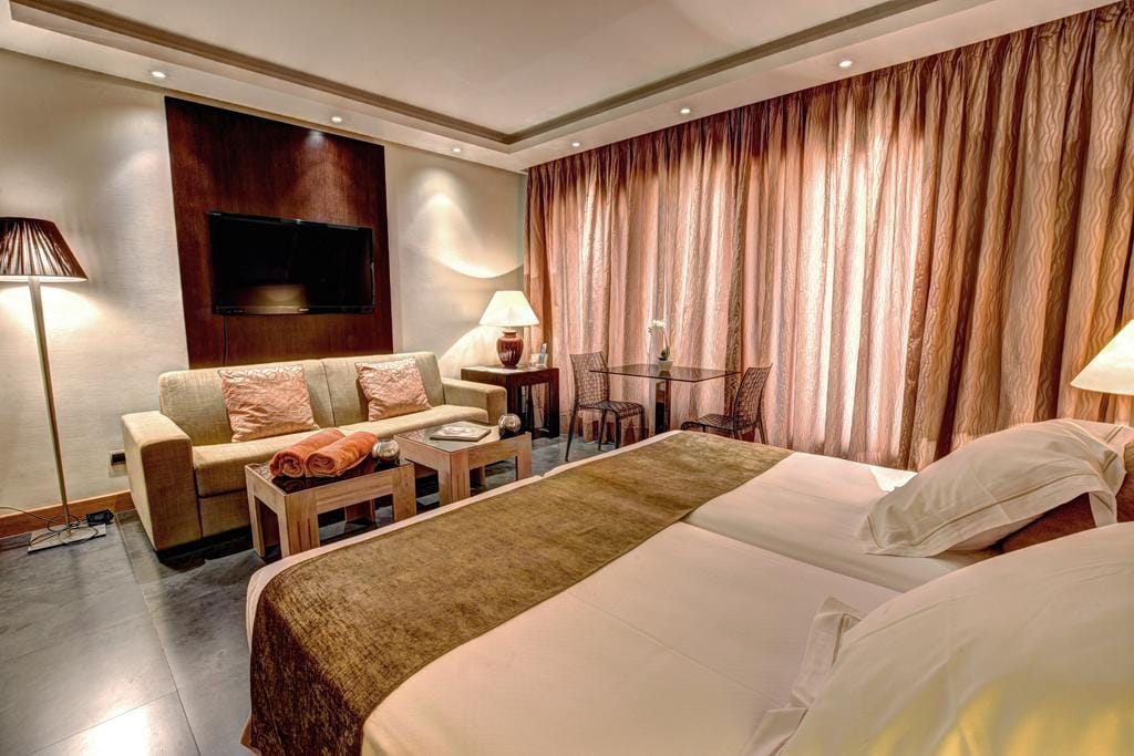 The spacious room features two individual King Size beds dressed in soft linens. A comfortable sofa and a small dining area are visible. A television is mounted on the wall, and soft lighting is provided by floor and table lamps, with floor-to-ceiling curtains allowing natural light.