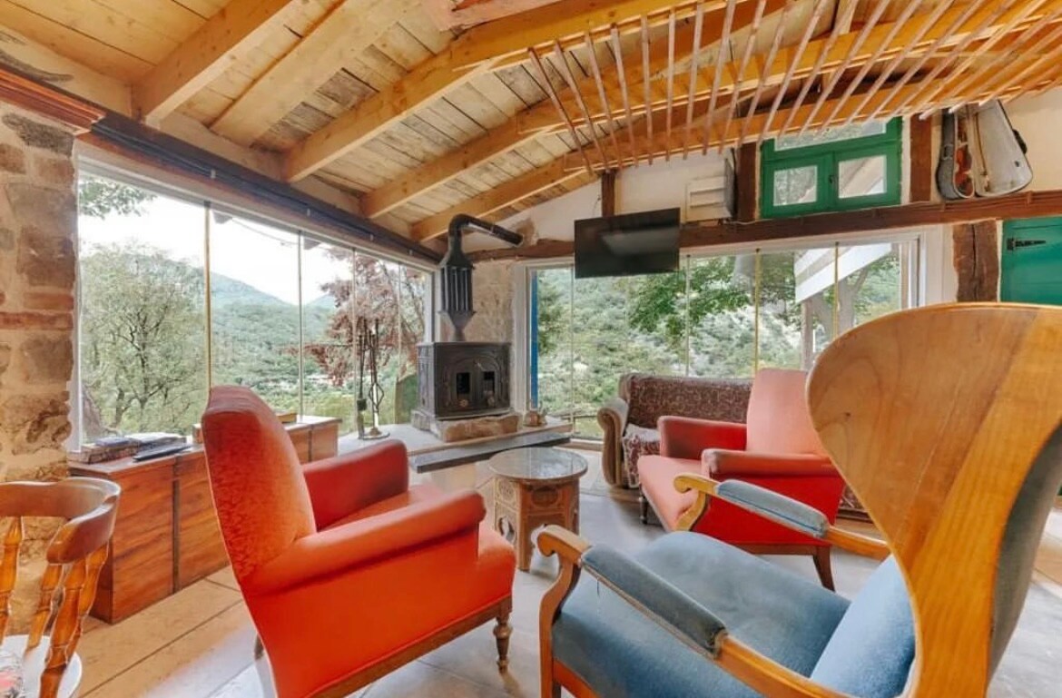 A rustic living area showcases a stone fireplace at its center, surrounded by an array of comfortable seating options in vibrant colors. Large windows offer views of the natural surroundings, allowing daylight to flood the space, while wooden beams add character to the ceiling.