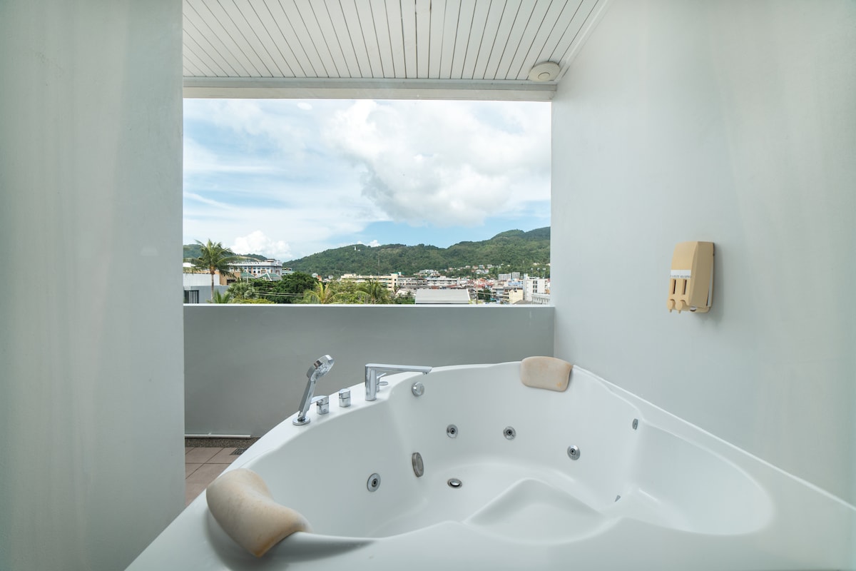 A spacious hot tub is positioned on a private balcony, offering an unobstructed view of the surrounding mountains and skyline. Natural light fills the area, creating a bright environment. The tub features multiple jets and soft edges for comfort.