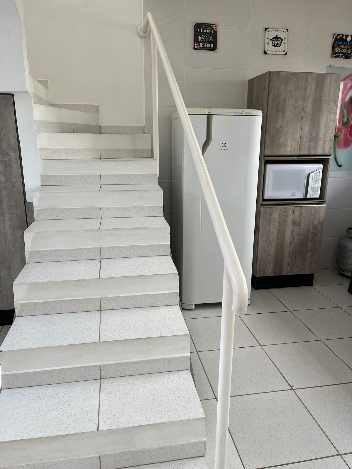 A white staircase leads to the upper level, flanked by a sleek white refrigerator and a compact microwave against a wooden cabinet. The tiled floor is designed in light and dark shades, enhancing the open space and ensuring safe navigation.