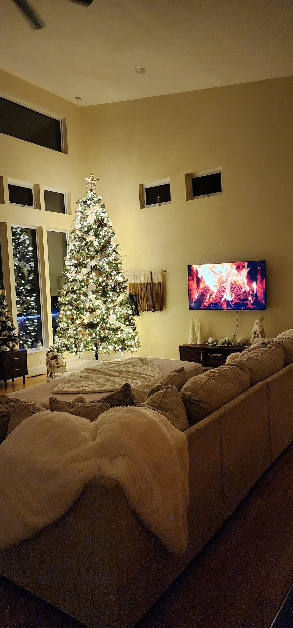A cozy living area features a large sectional sofa draped with a soft blanket. A beautifully decorated Christmas tree stands nearby, illuminated with warm lights. A television displays a flickering fireplace, enhancing the inviting ambiance of the space.