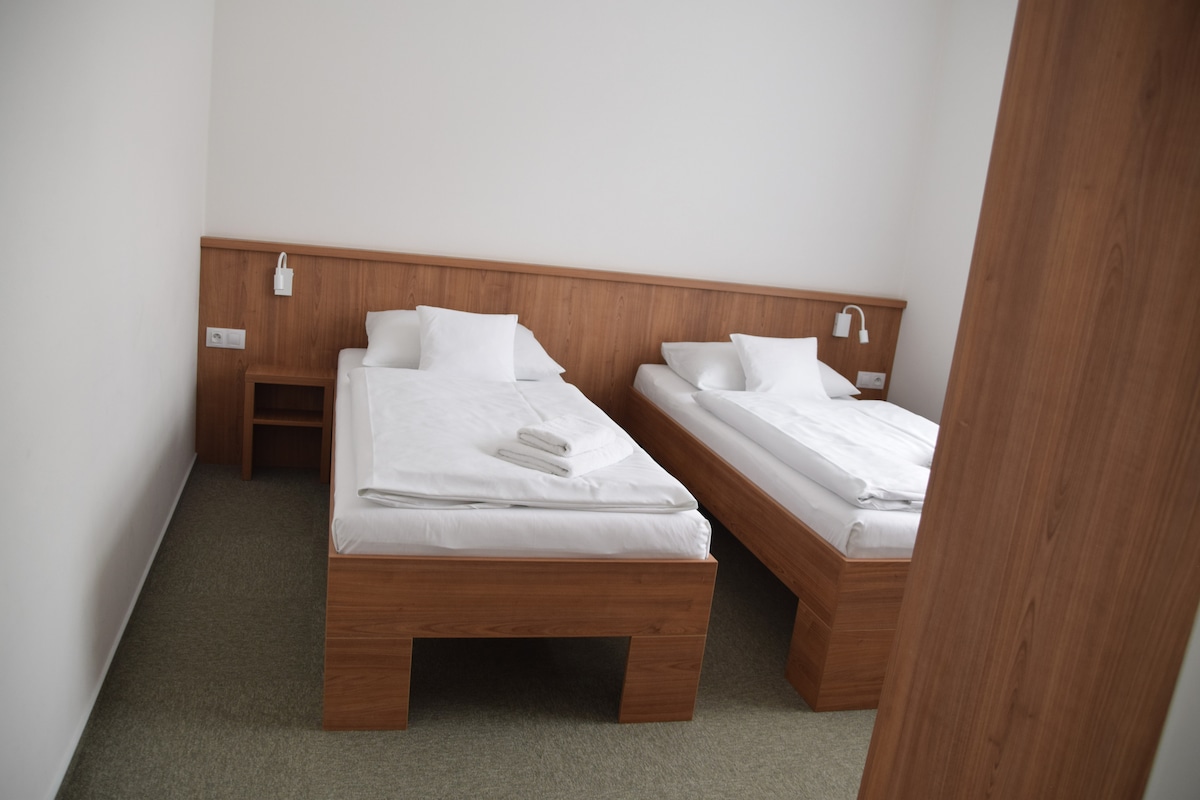 A simple room features two spacious beds with clean linens and neatly arranged towels on top. Light wooden headboards complement the minimalist design. Wall-mounted reading lights are installed above each bed, providing convenience for guests.