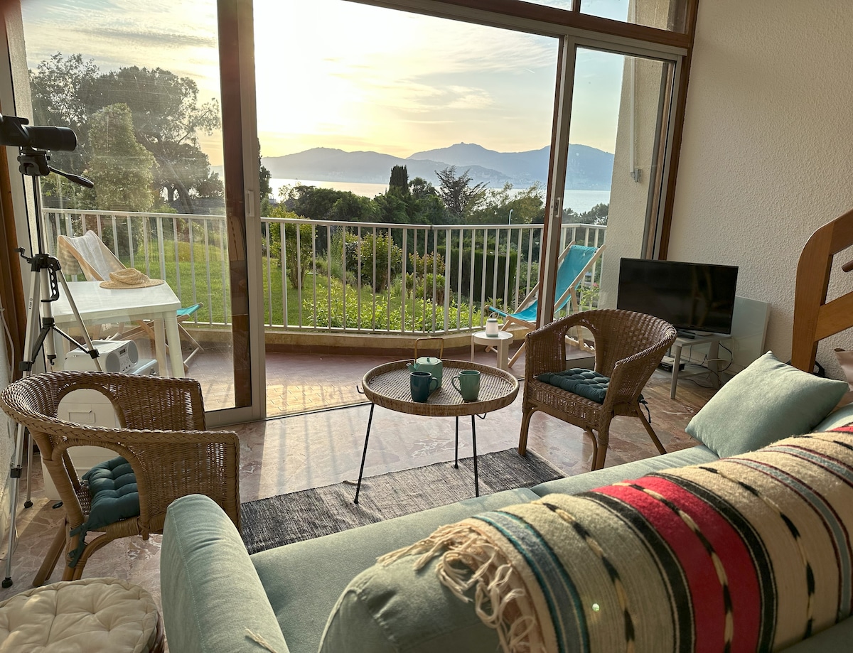 A bright living area is illuminated by soft morning light, featuring a comfortable sofa adorned with a striped throw. There are two wicker chairs and a small table with two cups, all positioned by large glass doors that open to a balcony with sea views.