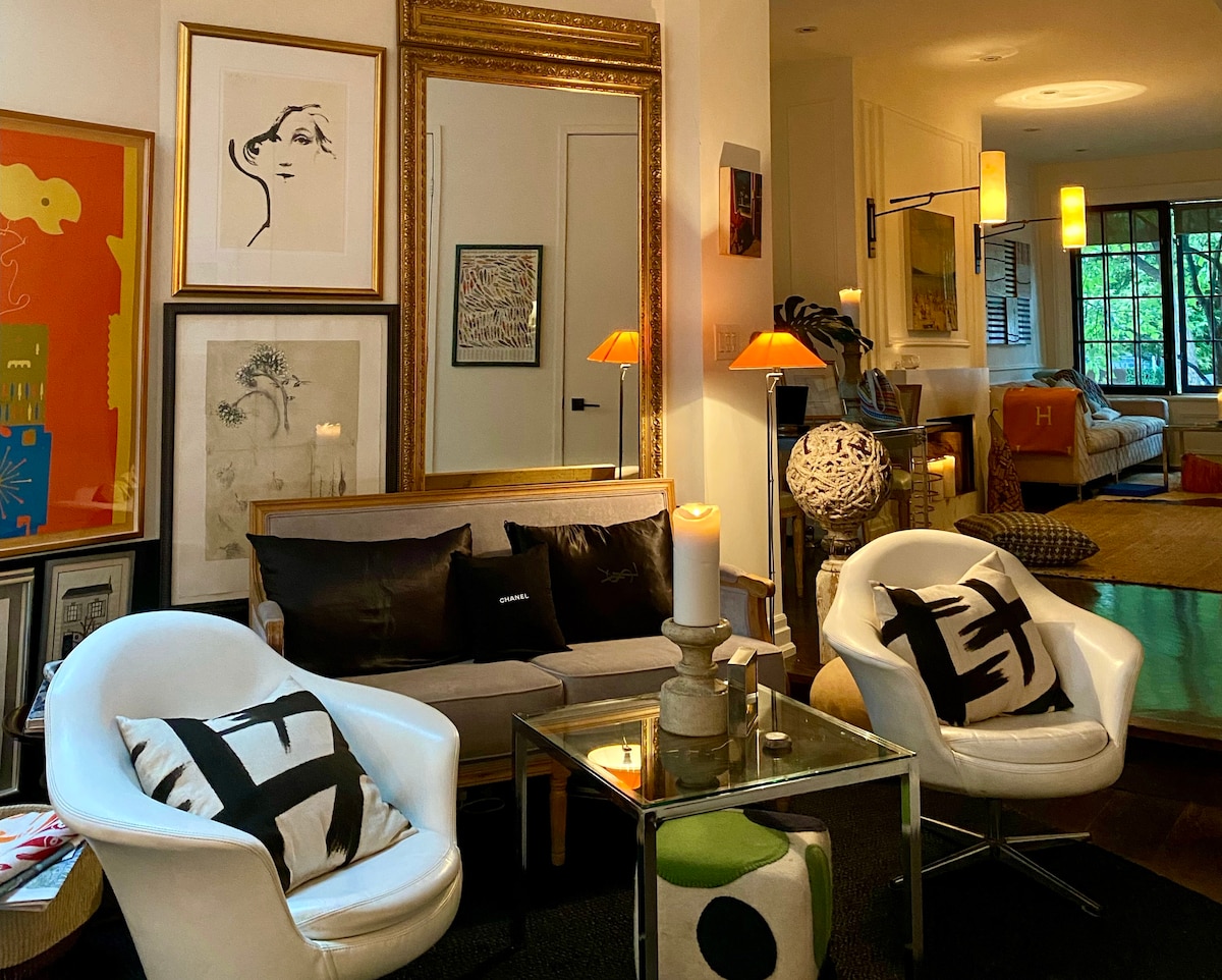 A welcoming living area features a stylish sofa accompanied by two white armchairs. Artworks in various frames adorn the walls, complemented by a decorative mirror. A glass coffee table with unique decor pieces sits at the center, illuminated by warm lamp lighting, creating an inviting ambiance.