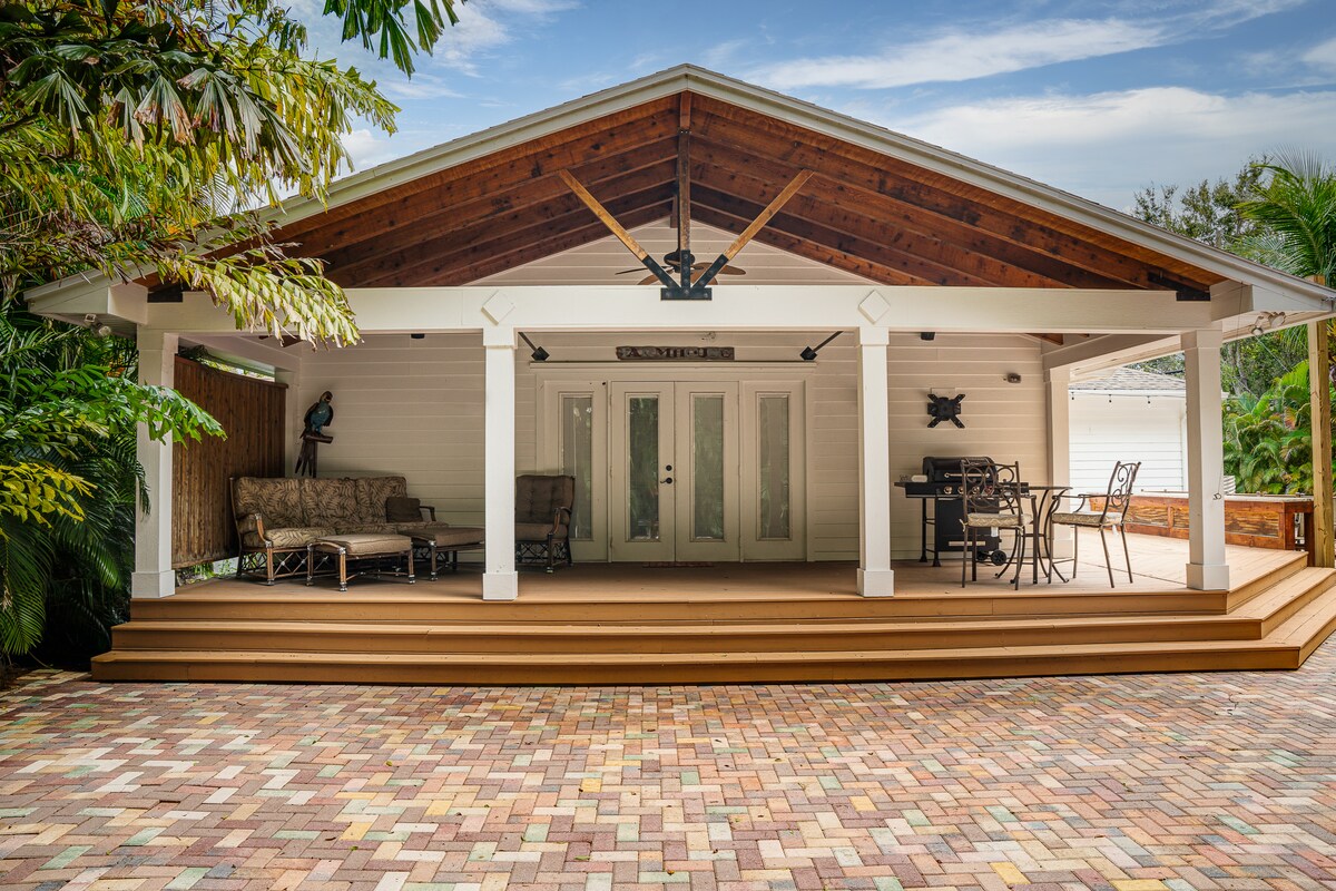 A spacious covered patio is framed by a high, wooden ceiling supported by beams. Comfortable seating and a dining area with a grill are visible, surrounded by lush tropical plants, creating an inviting outdoor space.