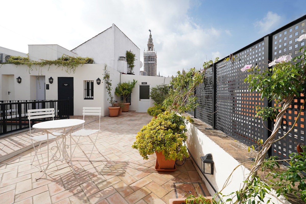 An outdoor terrace features a compact white metal table and two chairs, surrounded by potted plants and climbing greenery. The space provides a clear view of a historic tower in the background, under a cloudy sky with hints of blue.