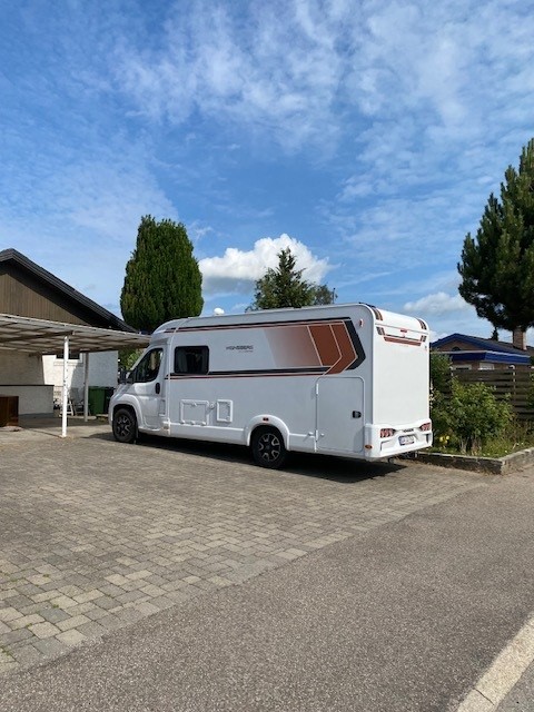 A modern camper van is parked on a paved area, featuring a sleek white exterior with orange accents. Surrounding greenery and a residential structure are visible, while a clear sky provides a spacious backdrop.