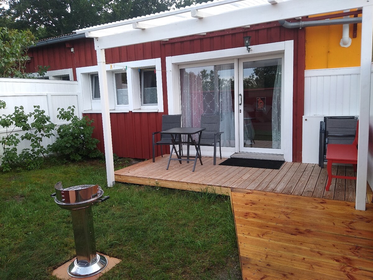 A boardwalk leads to a cozy outdoor seating area featuring a small table and two chairs, framed by green grass and shrubs. The entrance is highlighted by a glass door underneath a shaded patio, complemented by a grill positioned on the ground.