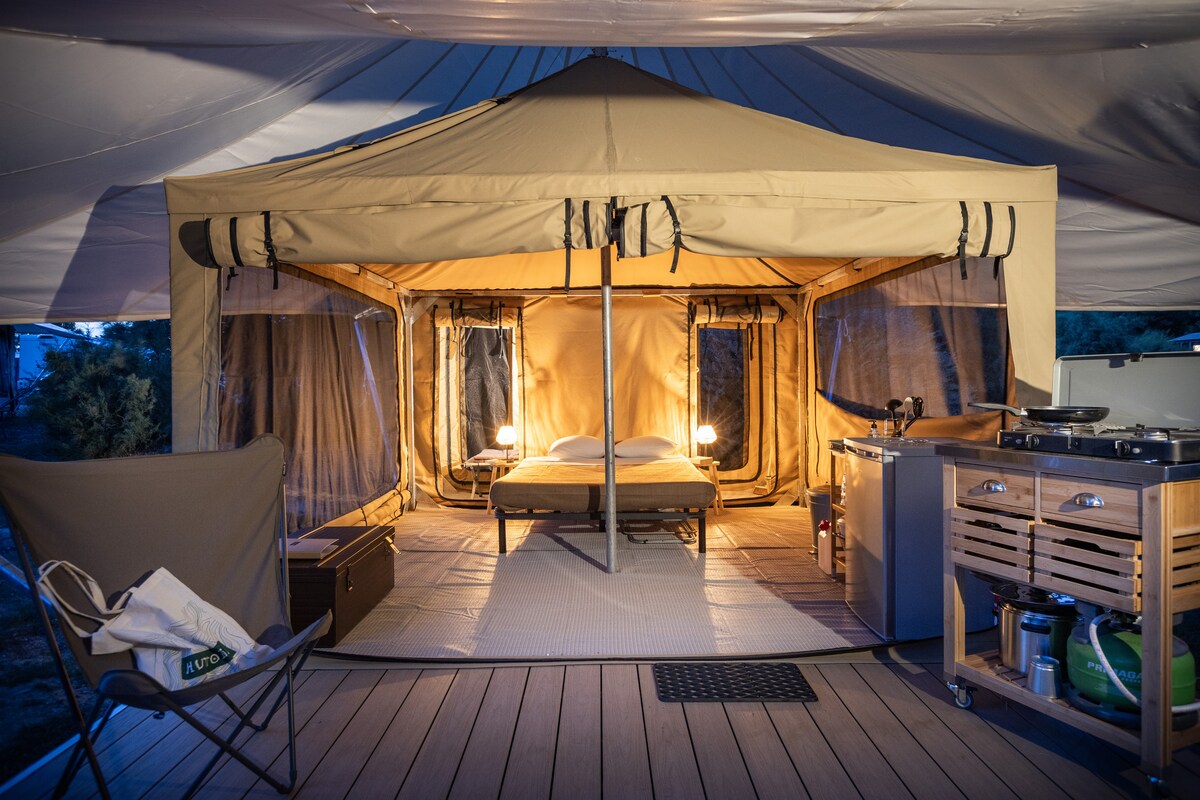 The tent interior features a spacious layout with a large double bed positioned centrally. Warm lighting from two bedside lamps creates a welcoming ambiance. A kitchenette is discreetly integrated, and the area is surrounded by large windows offering views of the outside environment.