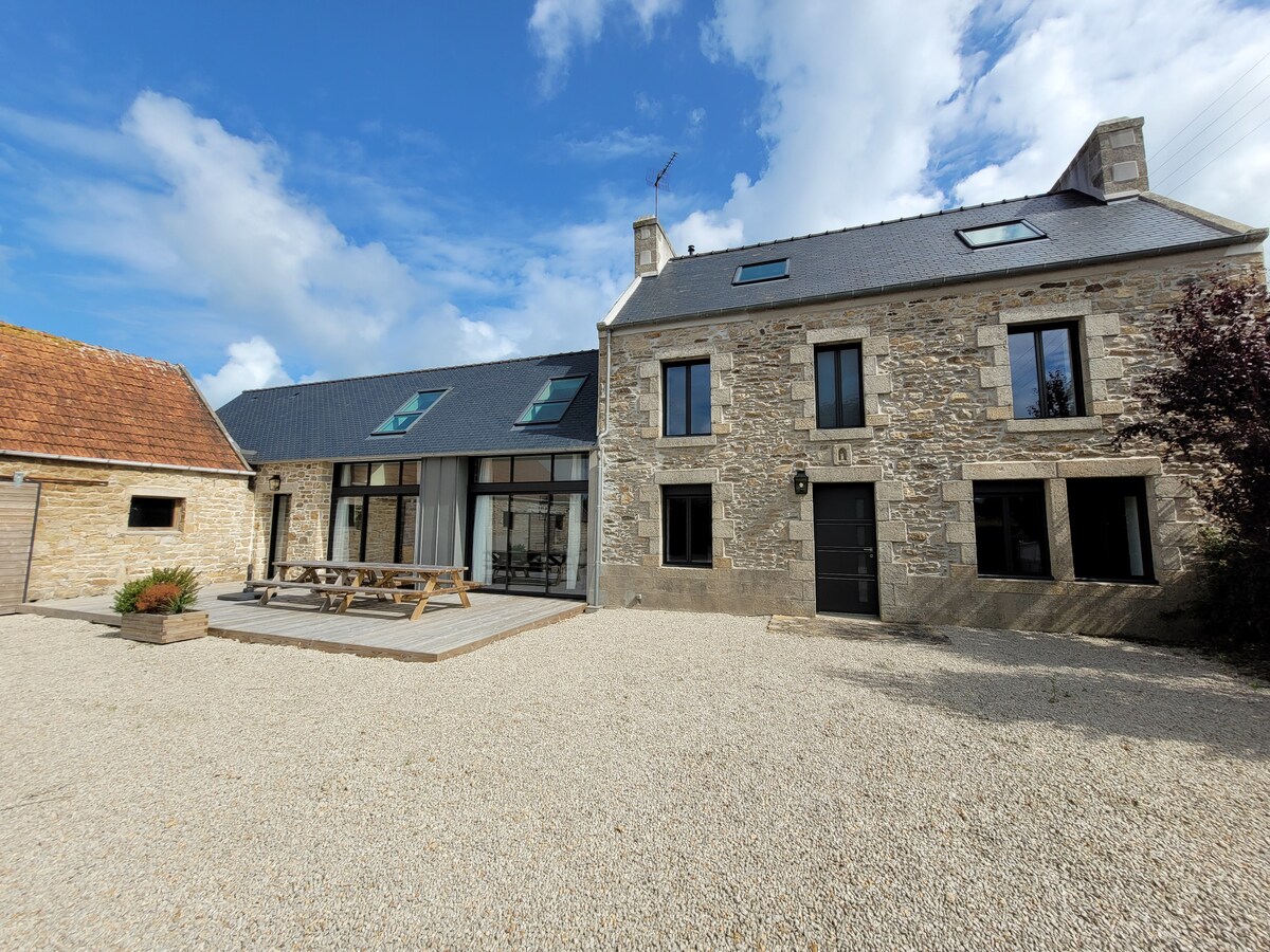 The exterior of the house features a charming stone facade with large windows allowing natural light. A spacious gravel area is visible, containing a wooden dining table on a deck. The structure includes a sloped roof and is framed by lush greenery.