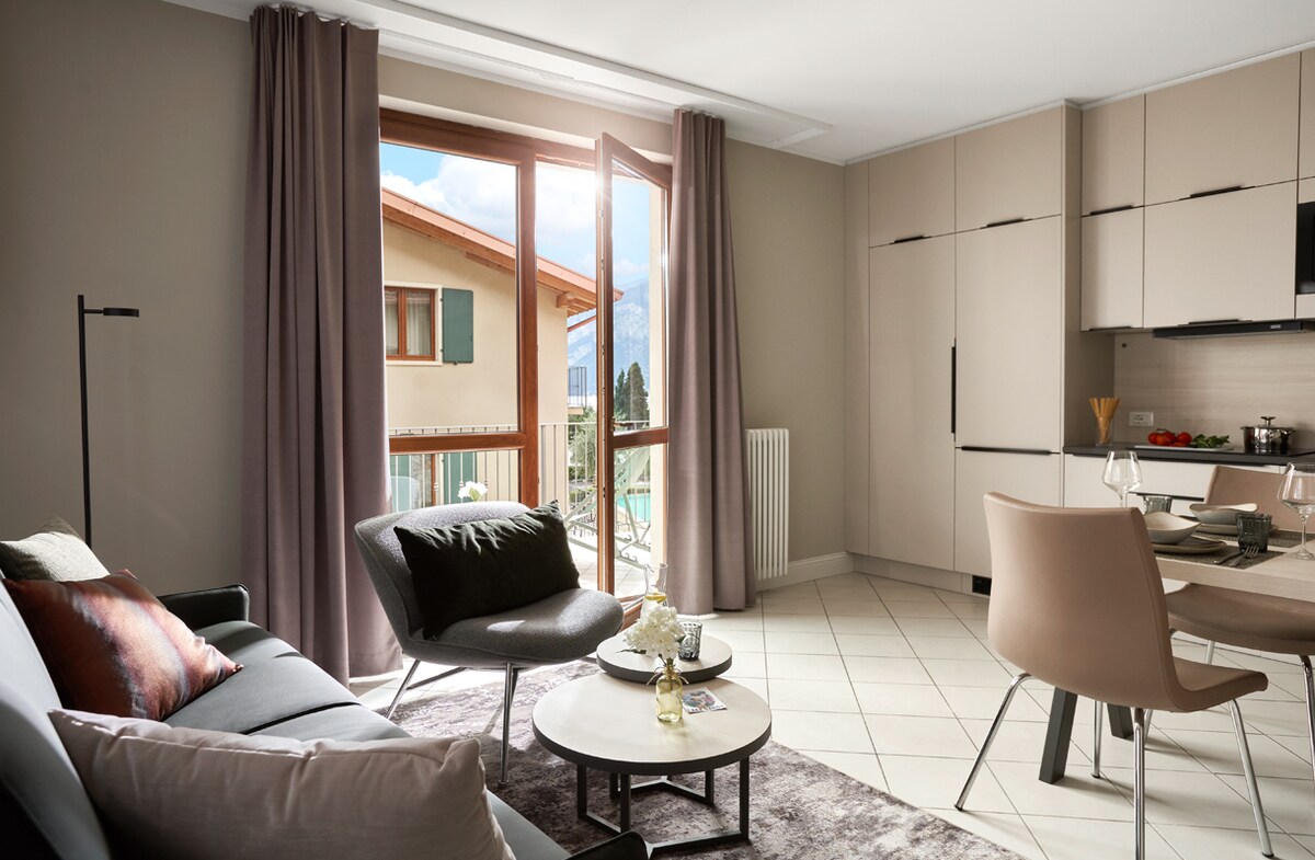 A bright living space features large open windows that provide side views of the lake and mountains. A comfortable sofa is paired with modern armchairs around a low coffee table. A dining area is visible, showcasing a light-colored table with elegant seating.