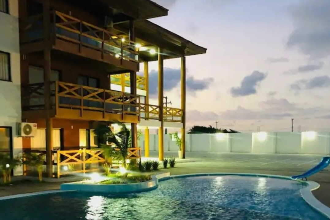 A modern building is visible beside a swimming pool, illuminated by soft lighting. The structure features multiple balconies with wooden railings and an outdoor area equipped with lounge chairs and palm plants. The scene is set against a twilight sky.