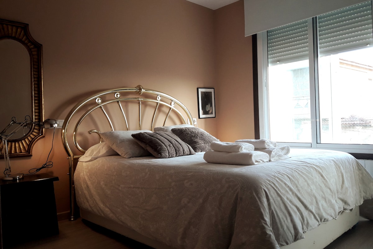 A spacious bedroom features a large bed with a decorative headboard and crisp linens. Pillows are neatly arranged on the bed, while a bedside table holds a lamp. Natural light fills the room through window blinds, and a framed art piece adds a touch of elegance.