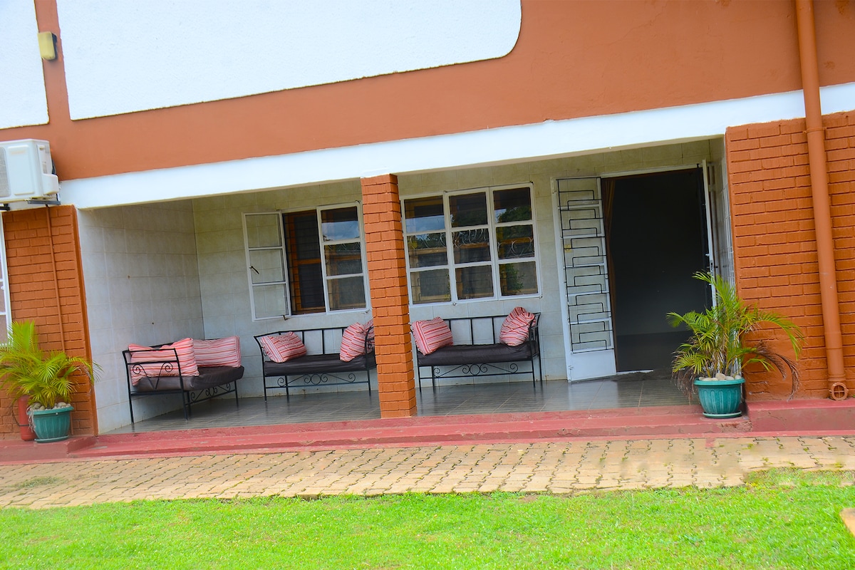 An outdoor seating area is presented with two black framed couches adorned with patterned cushions. Potted plants add a touch of greenery, while the entrance to the interior features a textured door. A spacious patio offers a smooth stone pathway leading into the garden.