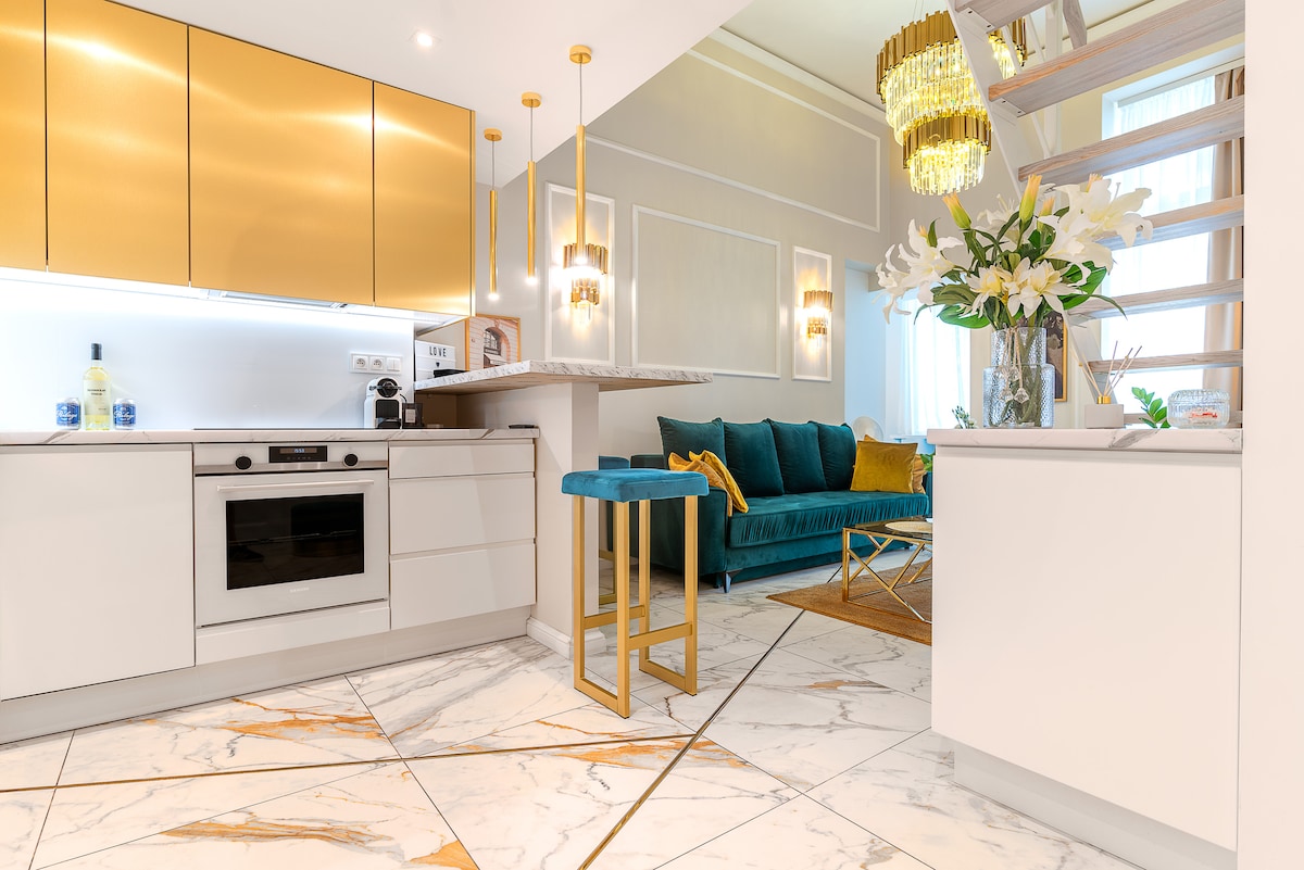 A modern kitchen area features sleek cabinetry with gold accents and state-of-the-art appliances. A spacious countertop separates the kitchen from a cozy living area, which includes a plush teal sofa and decorative pillows. The elegant marble flooring adds a touch of sophistication.