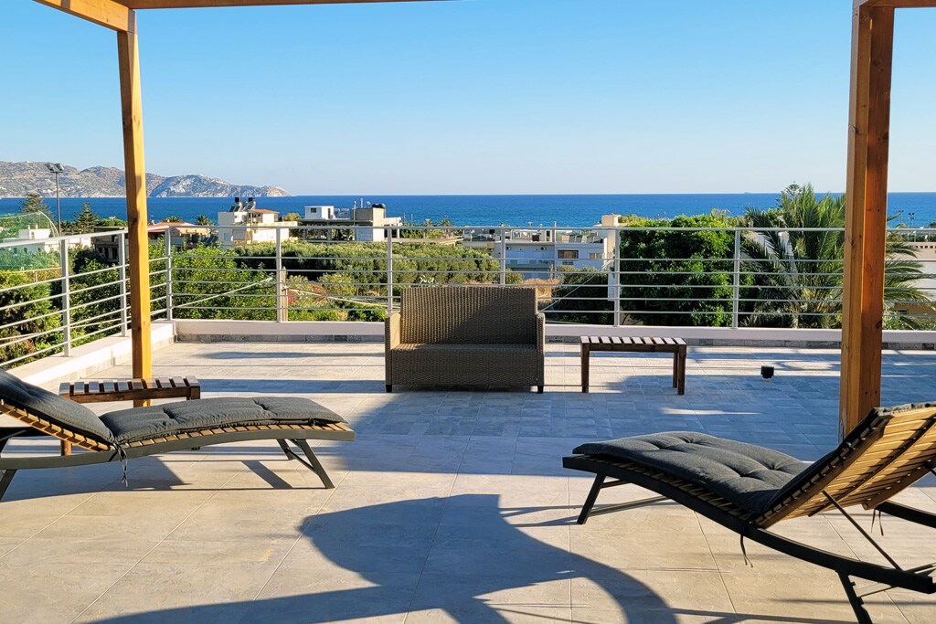 A spacious shared terrace is visible, featuring two reclining lounge chairs and a small wooden bench. The area offers a direct view of the sea and surrounding landscape, with clear blue skies above and lush greenery in the foreground.