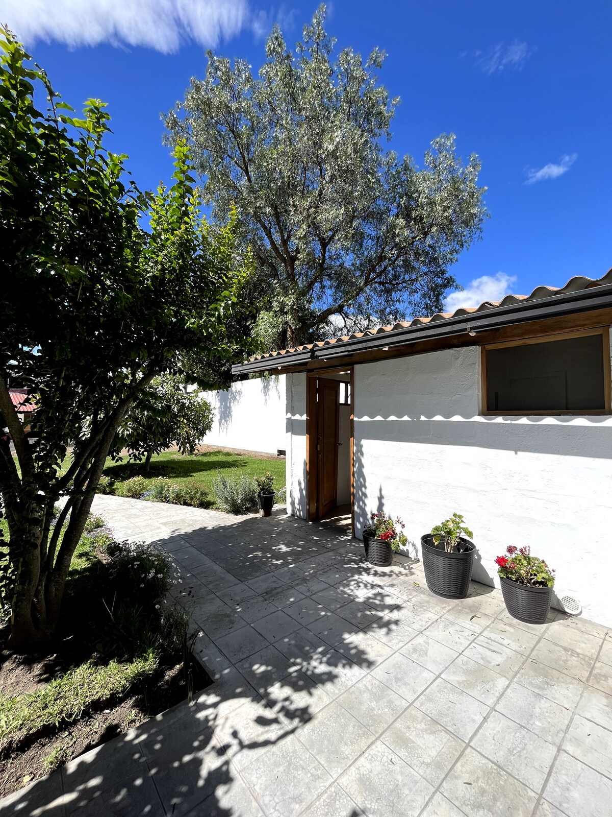 A pathway leads to a welcoming entrance adorned with potted flowers. A large tree provides shade, and a blue sky with scattered clouds enhances the serene outdoor setting. The entrance features wooden doors, inviting guests into a tranquil environment.