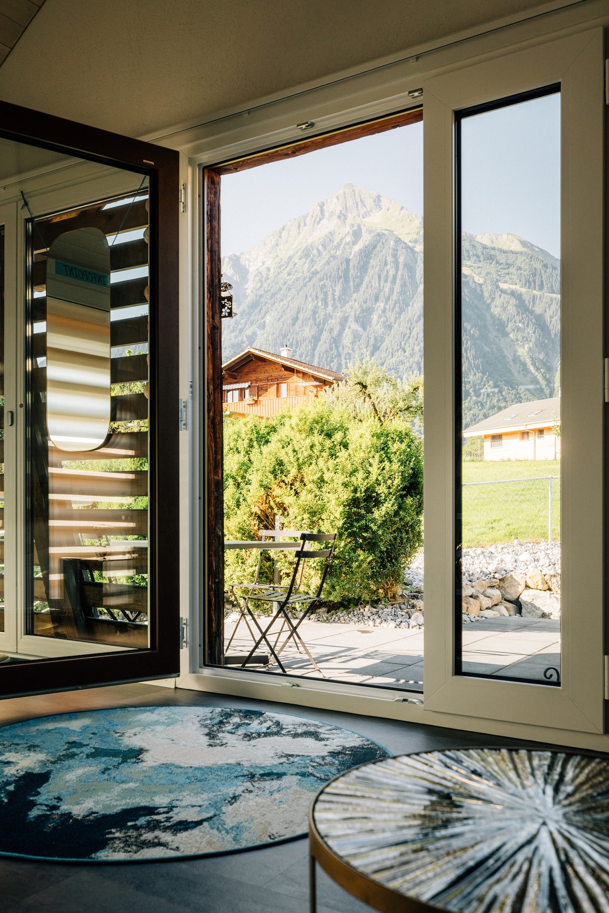 A view of the outdoor terrace is showcased through large glass doors, revealing a small table and two chairs set against a backdrop of mountains. Lush greenery and wooden structures are visible in the distance, enhancing the serene environment.
