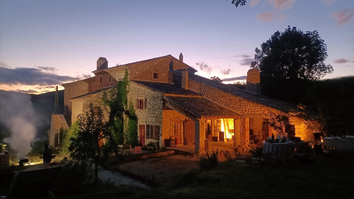 The stone house is illuminated by warm lights during twilight, highlighting its rustic architecture. Surrounding trees and foliage are visible, providing a natural frame. The atmosphere is peaceful, with a gentle smoke rising from a nearby area, suggesting a cozy outdoor gathering.