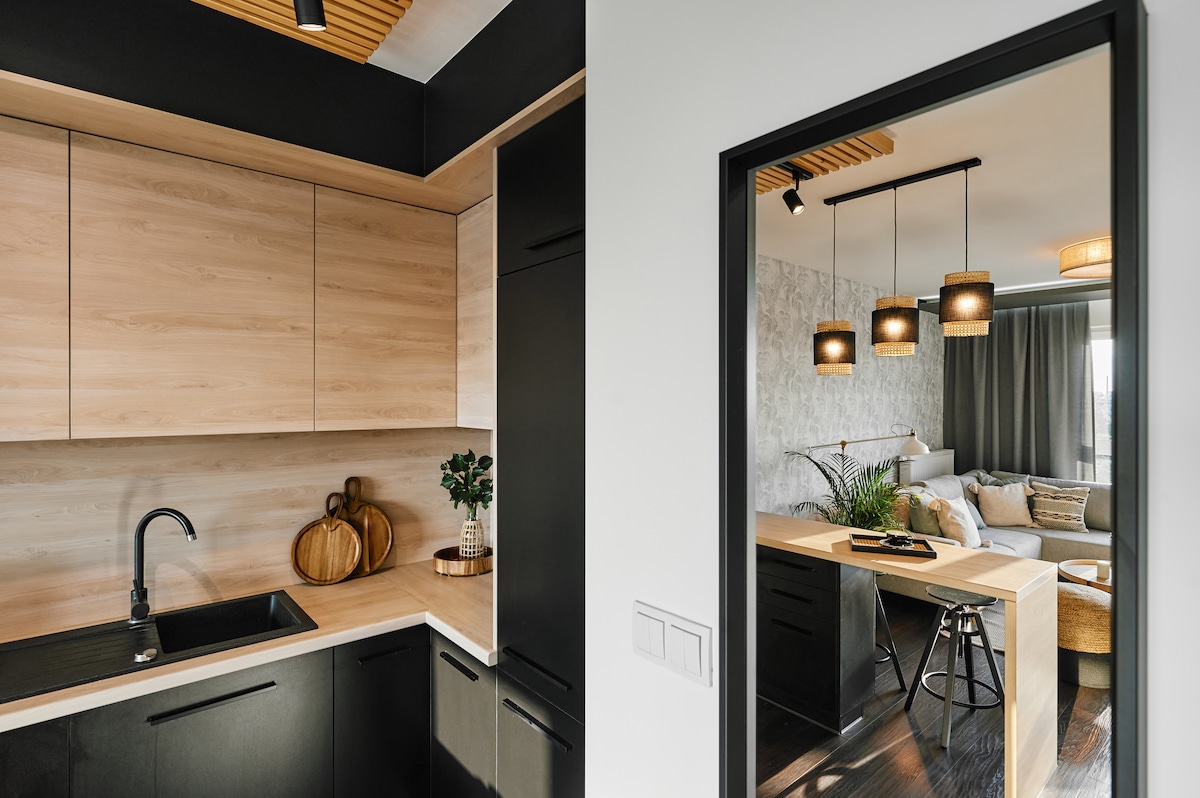 A well-equipped kitchen features cabinets in a combination of light wood and black finishes. A sleek black sink is positioned beside decorative items. A doorway offers a view of the inviting living area, highlighted by pendant lights and modern furnishings.