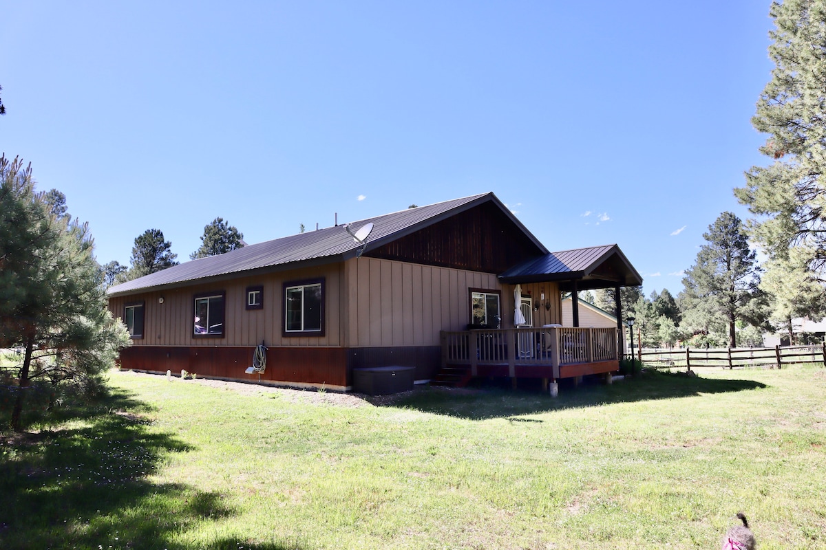 A spacious mountain home is seen from the exterior, highlighted by a large porch with a overhanging roof. The surrounding yard features green grass and a natural landscape with trees. The structure reflects a blend of wood and siding, set against a clear blue sky.