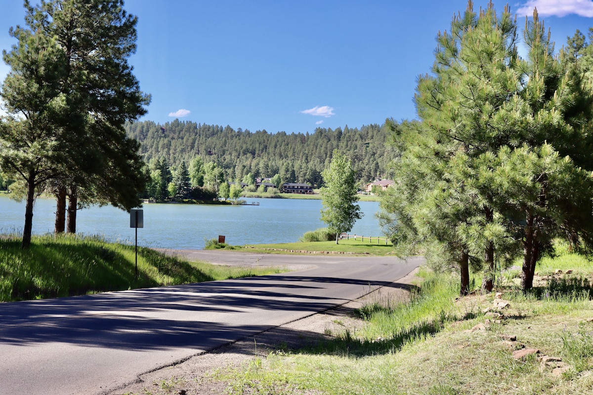 A serene view of a lake surrounded by lush green trees and gently rolling hills. The road leads towards the water, with clear blue skies reflecting the tranquil atmosphere. The natural surroundings suggest a peaceful outdoor retreat.