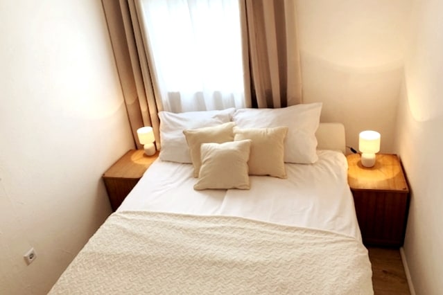 A well-lit bedroom features a spacious bed with a light-colored blanket and several pillows. Nightstands with soft lamps are placed on either side, creating a balanced and calm ambiance. Natural light filters in through sheer curtains, enhancing the serene environment.